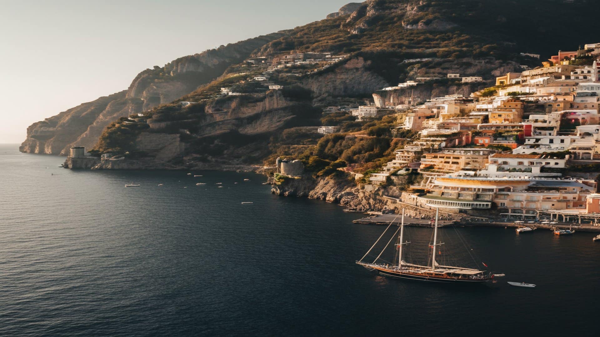 A European Mediterranean cliffside village at golden hour — pastel villas terraced above deep navy water, a single sailing yacht anchored offshore.